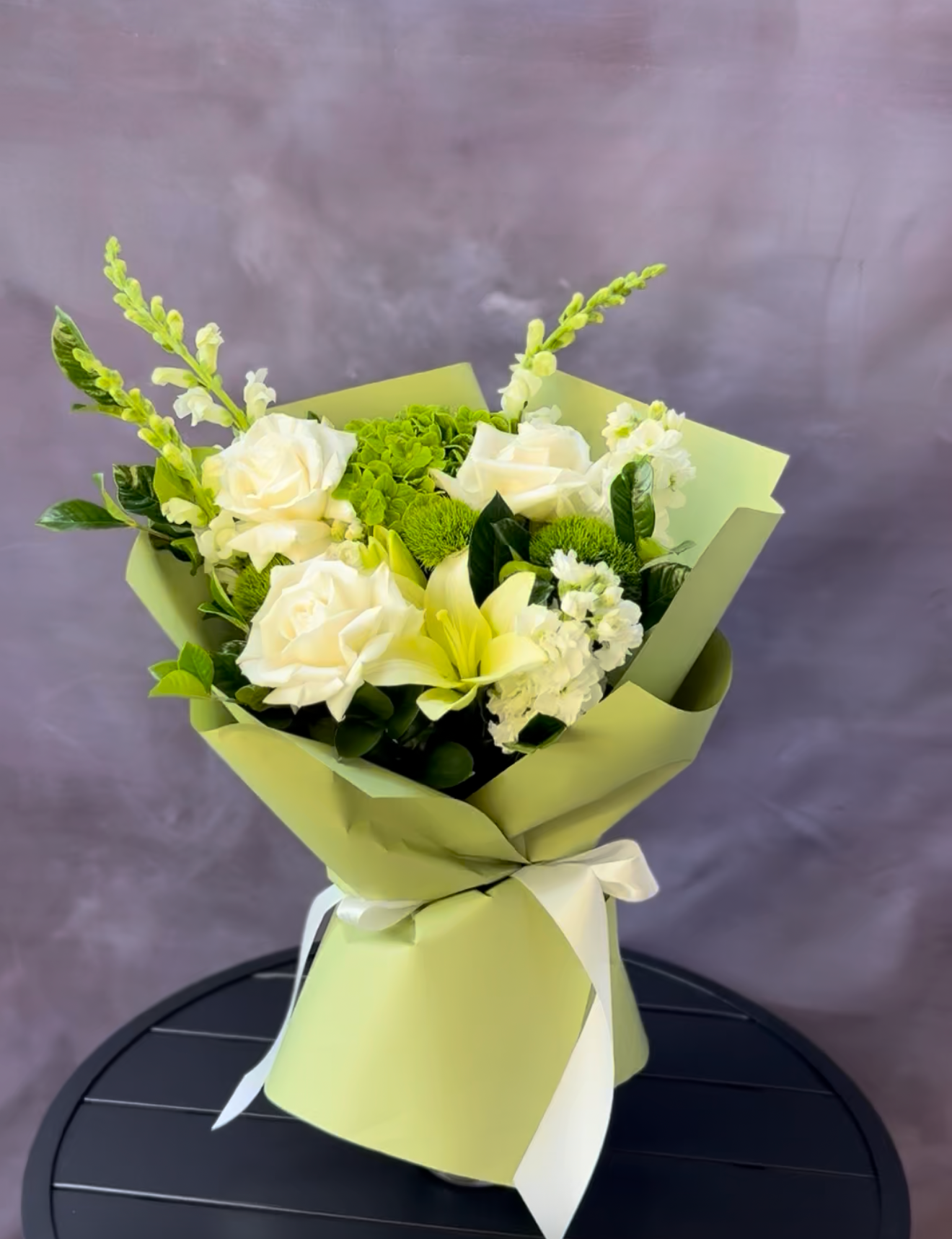 Bouquet of white and green flowers wrapped in green paper on a dark surface with a purple background